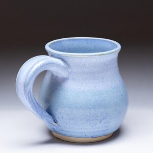 May include: A handmade ceramic mug with a light blue glaze. The mug has a rounded body, a curved handle, and a slightly flared rim. The base is unglazed, revealing the natural clay color. This mug is perfect for enjoying coffee or tea.