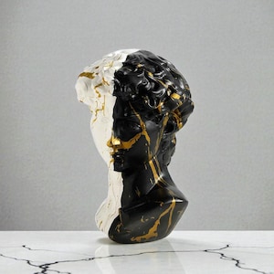 May include: A decorative bust of a classical figure, split vertically with a black and white color scheme. Gold paint accents the face and hair, creating a striking contrast. The sculpture is displayed on a white surface.