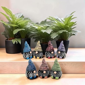 Puede incluir: Una colección de figuras de gnomos coloridos con cuerpos a rayas y sombreros puntiagudos en azul, amarillo, rojo y morado. Los gnomos están dispuestos sobre una superficie de madera frente a plantas verdes en macetas.