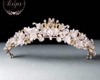 Könnte beinhalten: Ein goldfarbenes Diadem mit weißen blütenförmigen Akzenten, klaren Kristallen und kleinen weißen Perlen. Das Kopfstück hat ein symmetrisches Design mit einer zentralen Spitze und geschwungenen Seiten, die filigrane Metallarbeit zeigen.