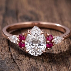 Könnte beinhalten: Ein Ring aus Roségold mit einem zentralen, birnenförmigen, klaren Edelstein, flankiert von zwei Gruppen aus je drei kleinen, runden, roten Rubinen und zwei äußeren, birnenförmigen, klaren Edelsteinen. Die Ringschiene ist glatt und poliert.