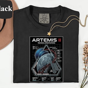 Puede incluir: Camiseta negra de cuello redondo con un gráfico de las especificaciones técnicas de la misión Artemis II. El diseño incluye una luna estilizada, la Tierra y formas geométricas con texto. Un collar con colgante de estrella dorada está sobre la camisa.