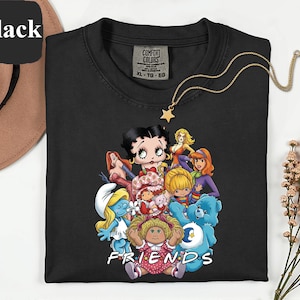 May include: Black Comfort Colors t-shirt featuring a graphic of cartoon characters including Betty Boop, Strawberry Shortcake, and others. The word "FRIENDS" is printed in white at the bottom. A gold star pendant hangs from a gold chain. A brown hat is in the background.
