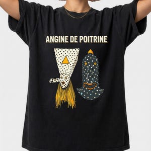 Könnte beinhalten: Schwarzes T-Shirt mit dem Text "ANGINE DE POITRINE" über zwei abstrakten Figuren. Eine Figur ist ein weißes Dreieck mit schwarzen Punkten und einem gelben Dreieck. Die andere ist eine schwarze Figur mit weißen Punkten.