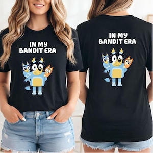 Könnte beinhalten: Schwarzes T-Shirt mit dem Text "IN MY BANDIT ERA" und einer Cartoonfigur auf der Vorder- und Rückseite. Die Cartoonfigur ist blau, gelb und orange. Das T-Shirt wird von einer Person getragen.