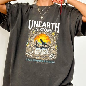 May include: Dark gray t-shirt with a graphic design featuring the text "UNEARTH A STORY" above a dinosaur scene. The design includes a dinosaur silhouette, an open book, and the text "2026 SUMMER READING".