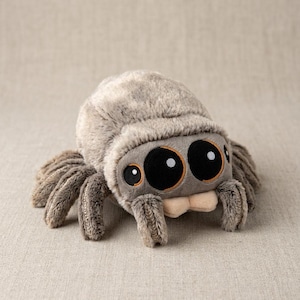 Puede incluir: Un peluche con forma de araña, de color gris y con grandes ojos negros. La araña de peluche tiene ocho patas y una boca beige. El juguete está diseñado con una textura suave, adecuado para niños.