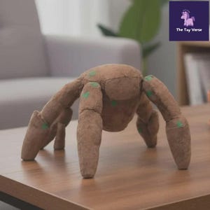 May include: A brown plush spider toy with eight articulated legs and green accents. The toy is made of a soft, textured material and is sitting on a wooden surface. The Toy Verse logo is in the background.