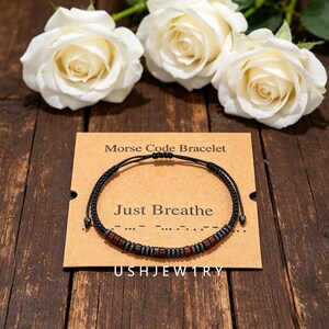 Könnte beinhalten: Ein schwarzes Schnurarmband mit abwechselnden dunkelbraunen Holzperlen und metallisch grauen Perlen in einem Morsecode-Muster. Das Armband liegt auf einer beigen Karte mit dem Aufdruck Morse Code Bracelet und Just Breathe.