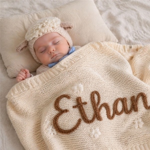 May include: A sleeping baby wearing a sheep hat and a blue bow tie, covered with a cream-colored knitted blanket. The blanket has the name "Ethan" embroidered in brown yarn, along with white flower accents. The baby is lying on a cream-colored pillow.