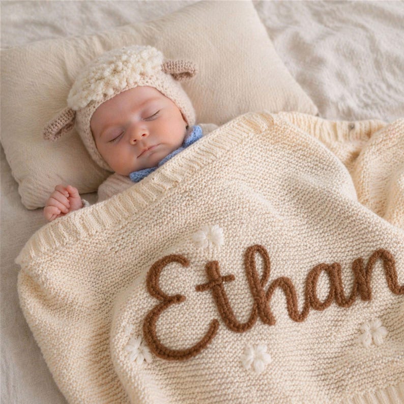 May include: A sleeping baby wearing a sheep hat and a blue bow tie, covered with a cream-colored knitted blanket. The blanket has the name "Ethan" embroidered in brown yarn, along with white flower accents. The baby is lying on a cream-colored pillow.