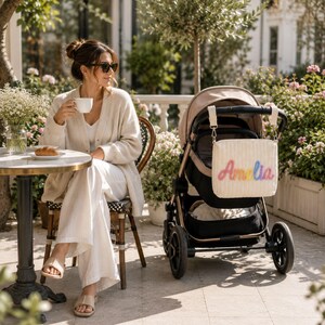 May include: A cream-colored, quilted rectangular stroller bag featuring the name Amelia embroidered in a vibrant, multi-colored gradient script. The bag is attached to the handlebar of a beige and black stroller with two white straps and clips.