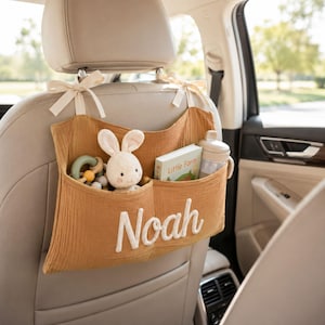May include: A mustard yellow fabric car seat organizer with two pockets, tied to the headrest with cream ribbons. The name Noah is embroidered in white script on the front. It holds a plush rabbit, a ring toy, and a book titled Little Farm.