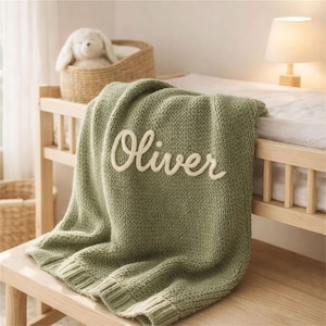May include: A sage green knitted baby blanket with the name "Oliver" embroidered in cream-colored script. The blanket is draped over a wooden changing table, with a stuffed bunny in a wicker basket in the background.