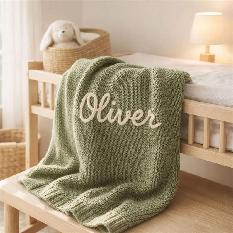 May include: A sage green knitted baby blanket with the name "Oliver" embroidered in cream-colored script. The blanket is draped over a wooden changing table, with a stuffed bunny in a wicker basket in the background.