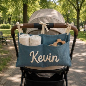 May include: A blue fabric stroller organizer featuring the name Kevin in white script lettering. The organizer is attached to the stroller handle with three cream-colored ribbons and contains two rolled white muslin cloths and a wooden toy.