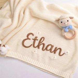 May include: Cream-colored knitted baby blanket with the name "Ethan" embroidered in brown. The blanket has a ribbed border and small flower accents. A lamb-shaped rattle with a blue bow is placed on the blanket.