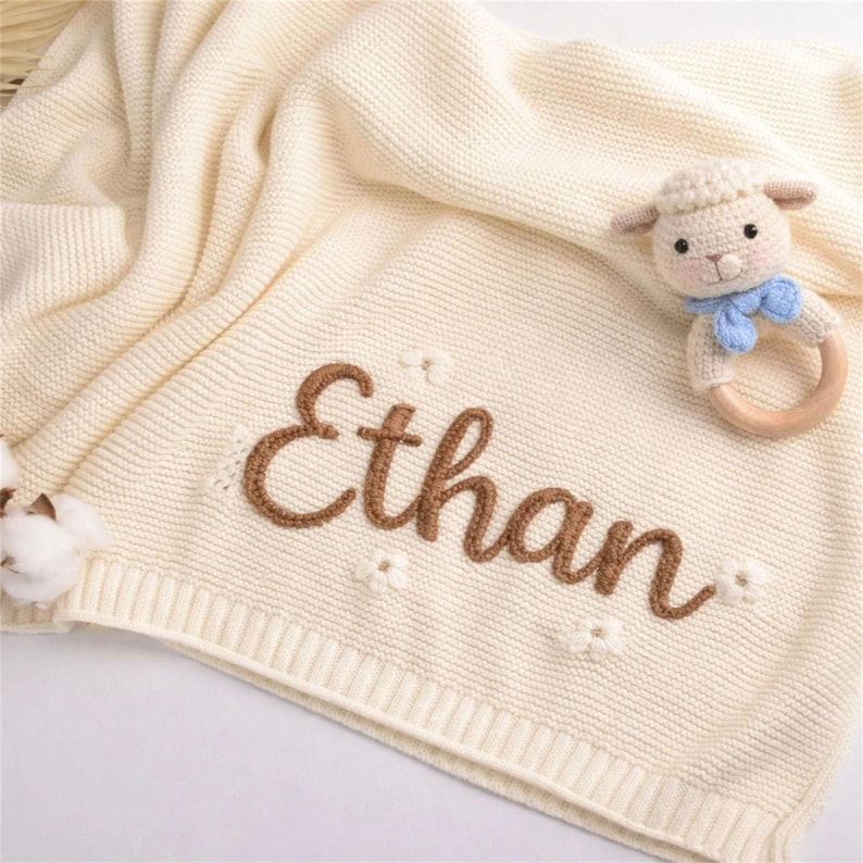 May include: Cream-colored knitted baby blanket with the name "Ethan" embroidered in brown. The blanket has a ribbed border and small flower accents. A lamb-shaped rattle with a blue bow is placed on the blanket.