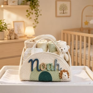 May include: A cream-colored rope storage basket featuring a rainbow design in shades of green and blue with the name Noah in dark blue and green script. The basket contains several folded light-colored cloths and a small plush bear toy.