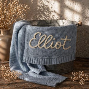 May include: A light blue knitted baby blanket with the name "Elliot" embroidered in cream-colored thread. The blanket is draped over a galvanized metal tub, with a portion of it resting on a wooden surface. Dried flowers are in a vase in the background.