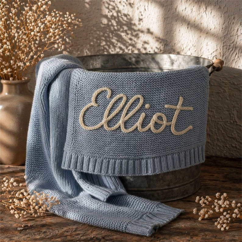 May include: A light blue knitted baby blanket with the name "Elliot" embroidered in cream-colored thread. The blanket is draped over a galvanized metal tub, with a portion of it resting on a wooden surface. Dried flowers are in a vase in the background.