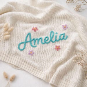 May include: Cream-colored knitted baby blanket with the name "Amelia" in turquoise script. The blanket is decorated with embroidered pink and purple flowers, creating a soft and inviting aesthetic. A cozy and personalized baby gift.