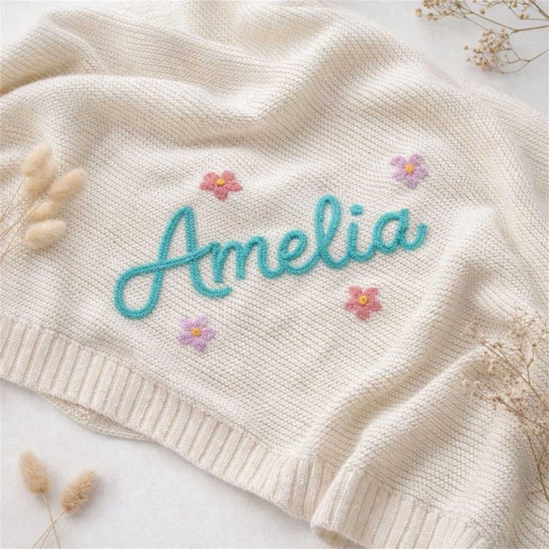 May include: Cream-colored knitted baby blanket with the name "Amelia" in turquoise script. The blanket is decorated with embroidered pink and purple flowers, creating a soft and inviting aesthetic. A cozy and personalized baby gift.