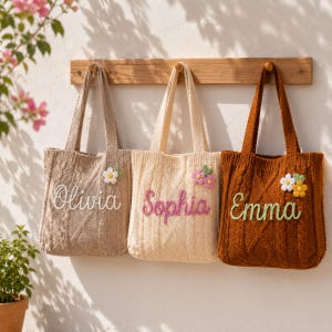 May include: Three cable-knit tote bags in beige, cream, and brown, each featuring a personalized name embroidered in cursive script and a small crocheted flower appliqué. The bags are suspended from a wooden rack mounted on a textured wall.