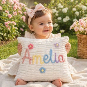 May include: A cream corduroy cushion embroidered with the name "Amelia" in colourful yarn. The cushion is decorated with small, embroidered flowers in pink, purple, and blue. A smiling baby holds the cushion.