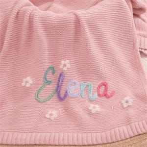 May include: A pink knitted blanket with the name "Elena" embroidered in colorful thread. The blanket is decorated with small white and pink flower appliqu&eacute;s, creating a soft and cozy aesthetic.