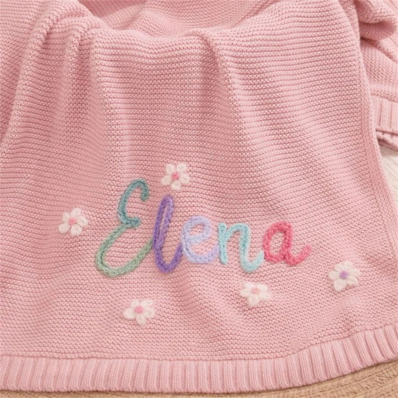 May include: A pink knitted blanket with the name "Elena" embroidered in colorful thread. The blanket is decorated with small white and pink flower appliqu&eacute;s, creating a soft and cozy aesthetic.