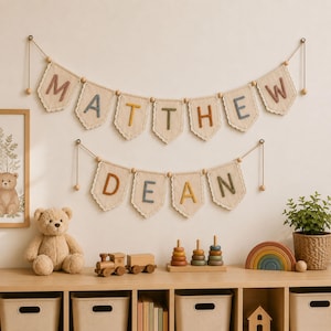 Könnte beinhalten: Ein Wandbanner-Set bestehend aus zwei Reihen von Stoffwimpeln mit Spitzenbesatz. Die obere Reihe buchstabiert MATTHEW und die untere Reihe DEAN in gestickten Buchstaben. Jeder Wimpel ist mit Holzperlen an dünnen Kordeln befestigt.