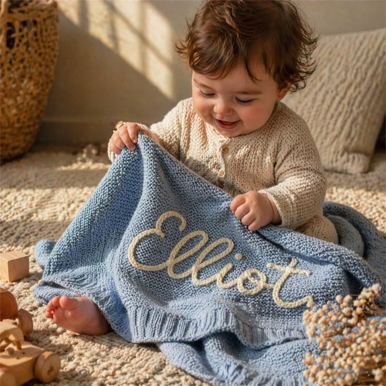 May include: A baby is holding a light blue knitted blanket with the name "Elliot" embroidered in cream-colored thread. The baby is wearing a beige knit outfit. The blanket is on a light-colored rug.