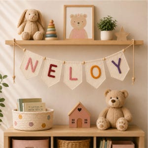Op de afbeelding: Een decoratieve stoffen slinger met vijf off-white wimpels opgehangen aan een touw met houten kralen. Elke wimpel toont een letter in een andere kleur, die samen de naam NELOY vormen in roze, donkerrood, roze, oranje en paars.