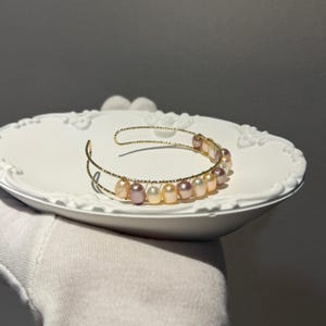 Könnte beinhalten: Ein goldfarbenes Armband in Manschettenform mit einem doppelt geführten, strukturierten Drahtband. Das Armband ist mit einer Reihe kleiner, ovaler Süßwasserperlen in verschiedenen Nuancen von zartem Rosa, Creme und Lavendel besetzt.