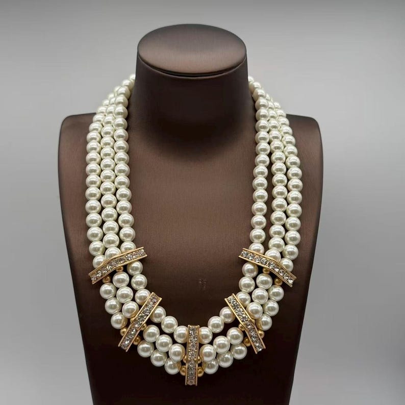 Peut inclure: Un collier &agrave; trois rangs compos&eacute; de perles blanches rondes dispos&eacute;es en lignes parall&egrave;les. Les rangs sont reli&eacute;s par cinq barrettes rectangulaires en m&eacute;tal dor&eacute;, chacune incrust&eacute;e d'une rang&eacute;e de petits cristaux transparents structur&eacute;s.