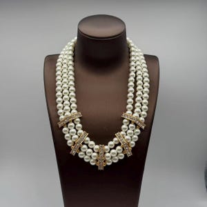 Peut inclure: Un collier &agrave; trois rangs compos&eacute; de perles blanches rondes. Les rangs sont reli&eacute;s par cinq barrettes rectangulaires en m&eacute;tal dor&eacute;, chacune incrust&eacute;e de petits cristaux transparents, cr&eacute;ant un aspect structur&eacute; et superpos&eacute;.