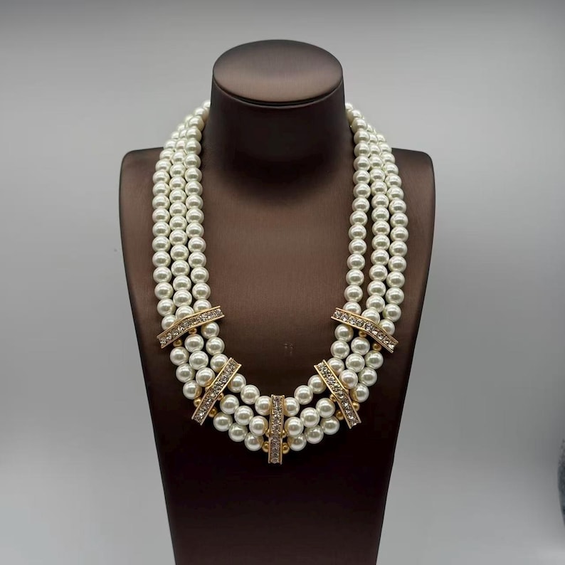 Peut inclure: Un collier &agrave; trois rangs compos&eacute; de perles blanches rondes. Les rangs sont reli&eacute;s par cinq barrettes rectangulaires en m&eacute;tal dor&eacute;, chacune incrust&eacute;e de petits cristaux transparents, cr&eacute;ant un aspect structur&eacute; et superpos&eacute;.