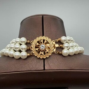 Peut inclure: Un collier multi-rangs compos&eacute; de trois rang&eacute;es de perles blanches sph&eacute;riques. Les rangs sont reli&eacute;s par un fermoir circulaire en m&eacute;tal dor&eacute;, orn&eacute; d'un motif g&eacute;om&eacute;trique complexe en forme de soleil avec un cristal transparent au centre.