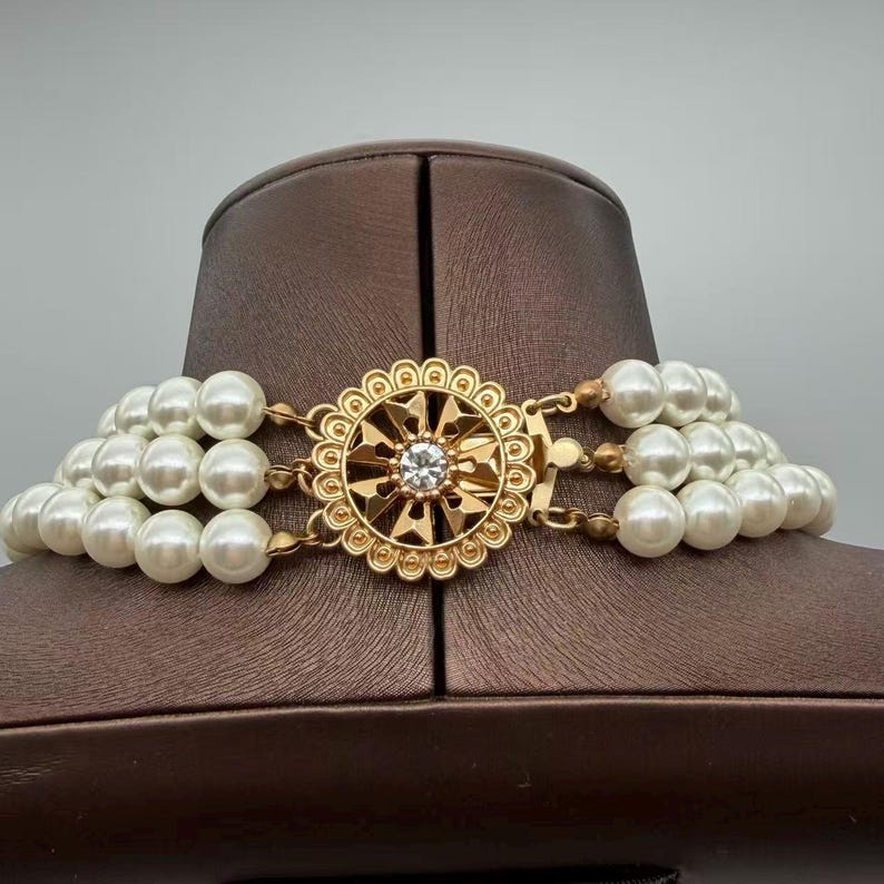 Peut inclure: Un collier multi-rangs compos&eacute; de trois rang&eacute;es de perles blanches sph&eacute;riques. Les rangs sont reli&eacute;s par un fermoir circulaire en m&eacute;tal dor&eacute;, orn&eacute; d'un motif g&eacute;om&eacute;trique complexe en forme de soleil avec un cristal transparent au centre.
