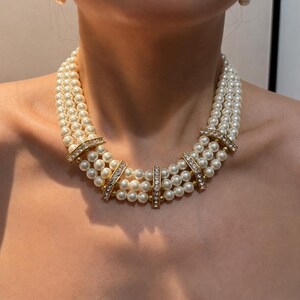 Peut inclure: Un collier &agrave; trois rangs compos&eacute; de perles blanches rondes. Les rangs sont maintenus par quatre entretoises rectangulaires en m&eacute;tal dor&eacute;, chacune orn&eacute;e d'une rang&eacute;e de petits cristaux transparents et &eacute;tincelants sur la partie avant.