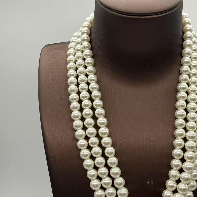 Peut inclure: Un collier multi-rangs compos&eacute; de trois rang&eacute;es distinctes de perles blanches, rondes et uniformes. Les perles pr&eacute;sentent une finition lisse et lustr&eacute;e, assembl&eacute;es dans un design continu et superpos&eacute; qui tombe verticalement sur le buste.