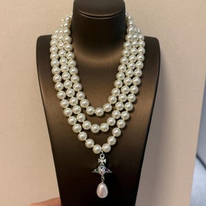 May include: A three-strand necklace featuring white round faux pearls of uniform size. The necklace includes a silver-toned metal orb pendant with colorful accents, topped with a cross, and finished with a single teardrop-shaped pearl dangling below.