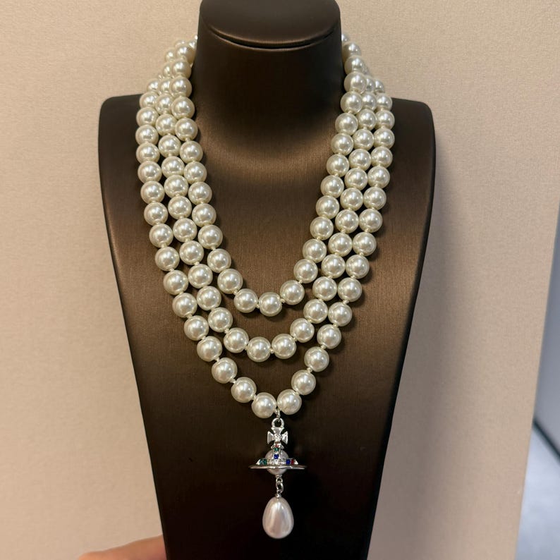 May include: A three-strand necklace featuring white round faux pearls of uniform size. The necklace includes a silver-toned metal orb pendant with colorful accents, topped with a cross, and finished with a single teardrop-shaped pearl dangling below.