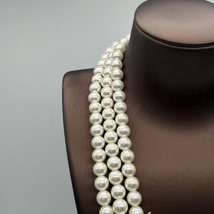 Peut inclure: Un collier multi-rangs compos&eacute; de trois rang&eacute;es de perles blanches, rondes et uniformes. Les perles pr&eacute;sentent une finition lisse et lustr&eacute;e, enfil&eacute;es en lignes parall&egrave;les pour cr&eacute;er un aspect superpos&eacute; qui tombe verticalement le long du cou.