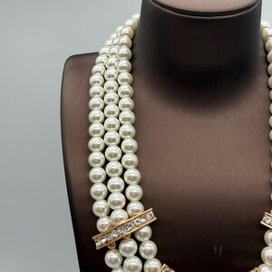 Peut inclure: Un collier multi-rangs compos&eacute; de trois rang&eacute;es de perles artificielles rondes, blanches et lustr&eacute;es. Les rangs sont reli&eacute;s par des entretoises rectangulaires en m&eacute;tal dor&eacute; incrust&eacute;es de petits cristaux clairs et &eacute;tincelants.