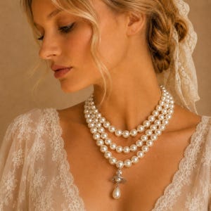 May include: A three-strand pearl necklace featuring round white pearls and a silver-toned orb pendant with a dangling teardrop pearl. The necklace is worn with a white lace garment that has a scalloped neckline and intricate floral embroidery.