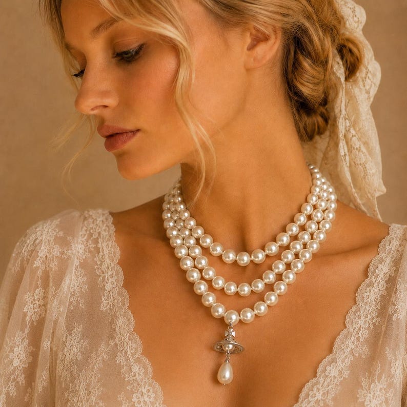 May include: A three-strand pearl necklace featuring round white pearls and a silver-toned orb pendant with a dangling teardrop pearl. The necklace is worn with a white lace garment that has a scalloped neckline and intricate floral embroidery.