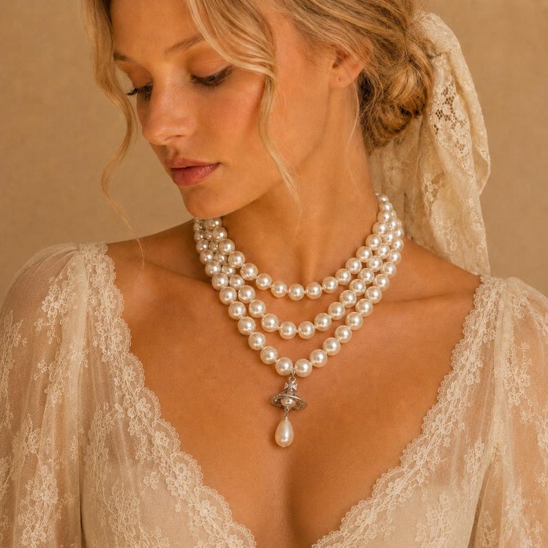 May include: A three-strand pearl necklace featuring round white pearls and a silver-toned orb pendant with a dangling teardrop pearl. The necklace is worn over a cream-colored lace garment with long sleeves and intricate floral embroidery details.