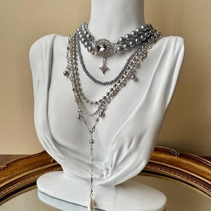 May include: A multi-strand necklace set, presented on a white bust. The set features silver-toned chains, beads, and faux pearls. A large, decorative pendant with a starburst design is suspended from the centre. The necklace is a statement piece.